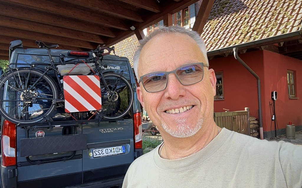 A smiling man taking a selfie in front of a camper van with a bicycle mounted on the back, under a wooden structure.