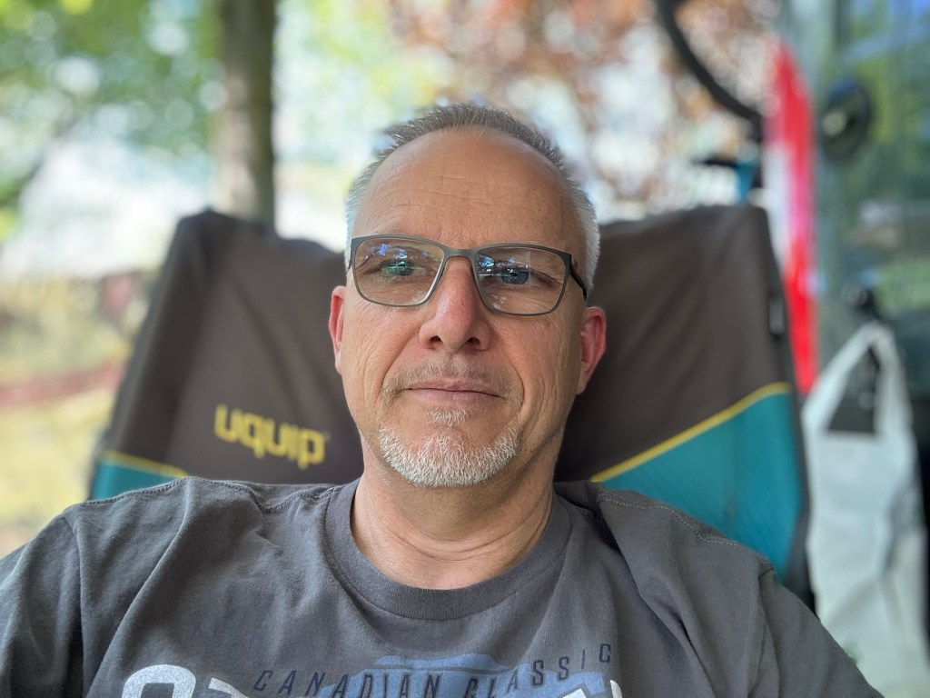 A person sitting in a camping chair with glasses, looking relaxed in a natural outdoor setting.