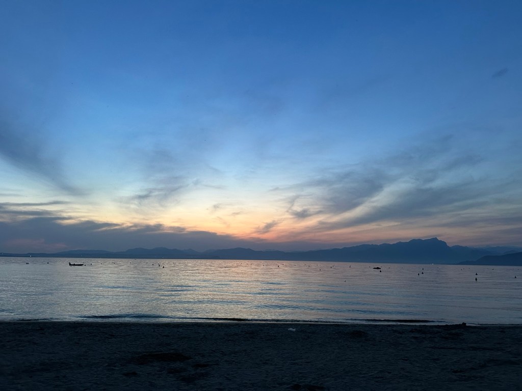 A serene sunset over Lake Garda, with gentle waves reflecting the orange and blue hues of the sky. The sandy shoreline is visible in the foreground, with distant mountains along the horizon.