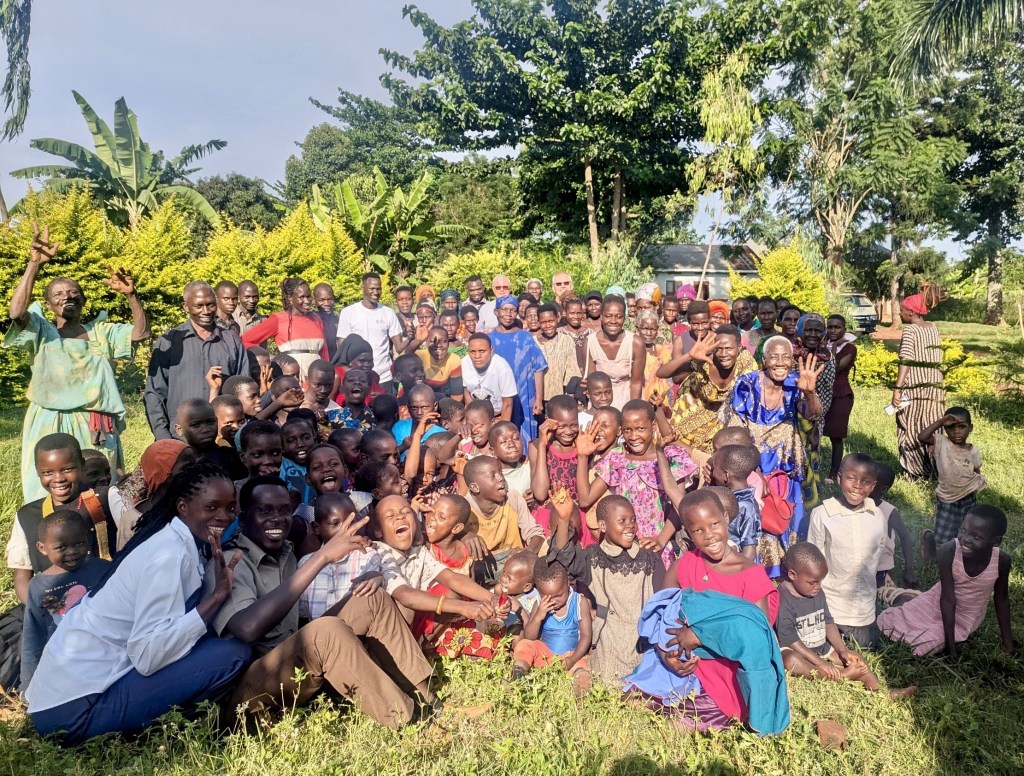 A large group of diverse men, women, and children gathered outdoors in a lush green environment, smiling and posing for the camera, with some waving and expressing joy.