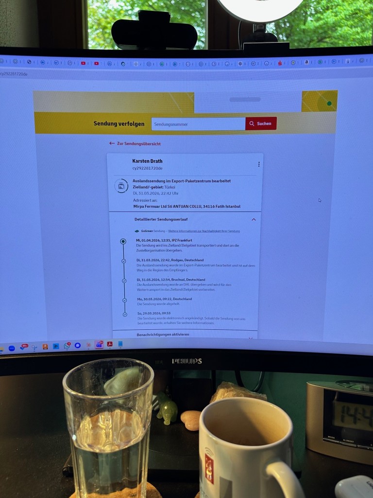 A computer monitor displaying a shipment tracking page with details including the sender and recipient information, status updates, and timestamps, alongside a glass of water and a mug on a desk.