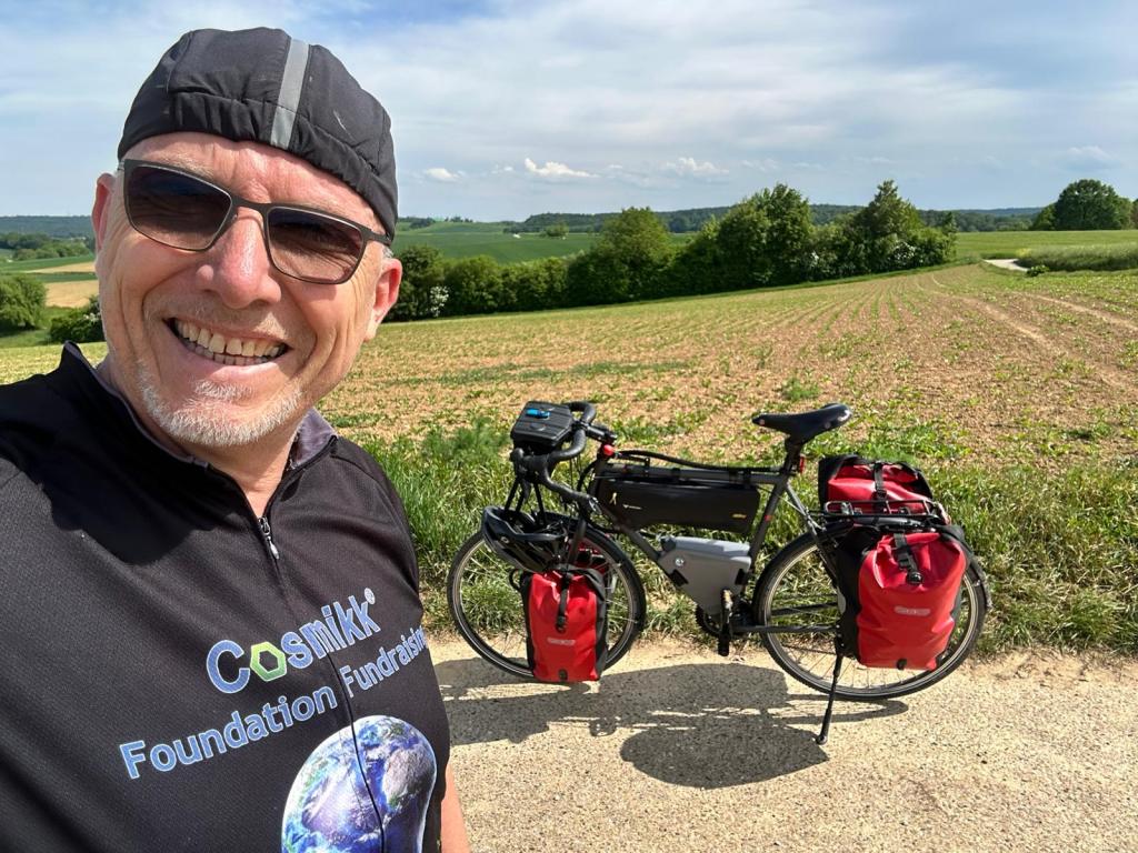 A smiling cyclist wearing sunglasses and a cycling jersey stands next to a bicycle with red panniers, set against a scenic landscape with fields and trees.